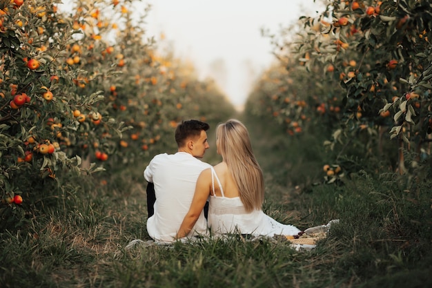 Beautiful couple dressed in elegant clothes sitting on green grass in apples orchard at summer picnic.