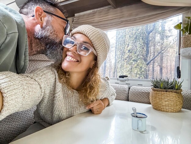 美しいカップルがキャンピングカーでキスをし携帯電話で写真を撮り微笑んでいます夫と妻は森で幸せに休暇を楽しんでいます