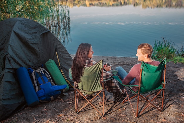 Beautiful couple sitting together on folding chairs at water and look at each other. They smile. She holds cup in hands and covered with blanket. There are tent and equipment as well.