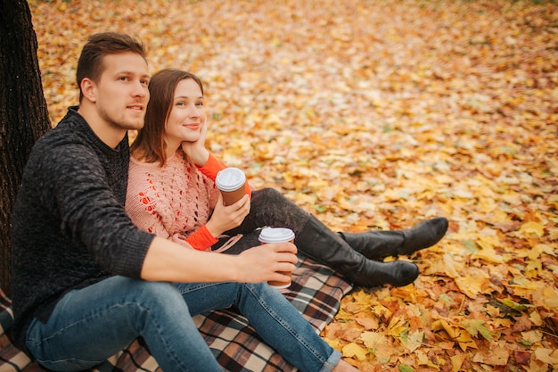 Beautiful couple sitting together and look in one direction. They hold cups of coffee. He embrace her. They sit in park.