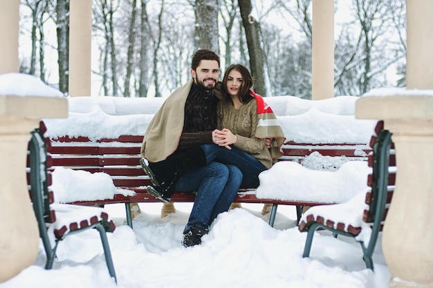 Beautiful couple in snowy park. young man, woman stay near snow covered pine trees in warm sweaters.