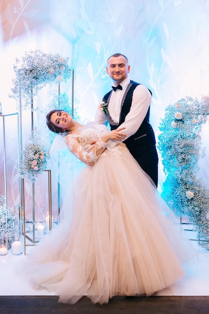 A beautiful couple stands against the background of the photo zone the bride bending over in a long dress a man in a tuxedo happy couple smiles