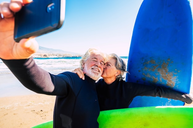 Beautiful couple of two seniors at the beach with wetsuits and surfboard taking a selfie before go surf together - active mature people