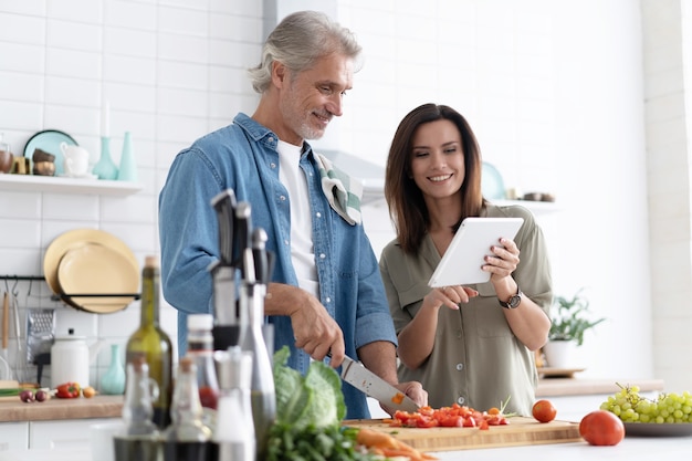 Красивая пара с помощью цифрового планшета и улыбается во время приготовления пищи на кухне дома