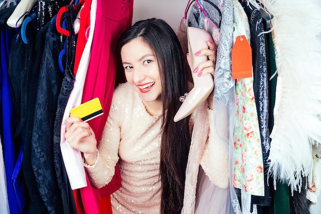 Beautiful crazy brunette mad woman shopaholic looks through the hangers wardrobe with clothes holding a shoes and a plastic credit card in her hand concept of seasonal sales and shopaholicism