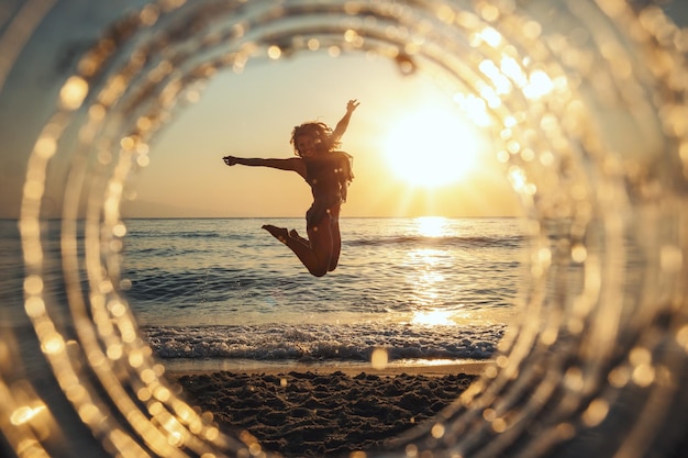 Una bella composizione creativa di un paesaggio marino girato attraverso una messa a fuoco circolare che mostra una giovane donna che sta saltando sulla spiaggia al tramonto.