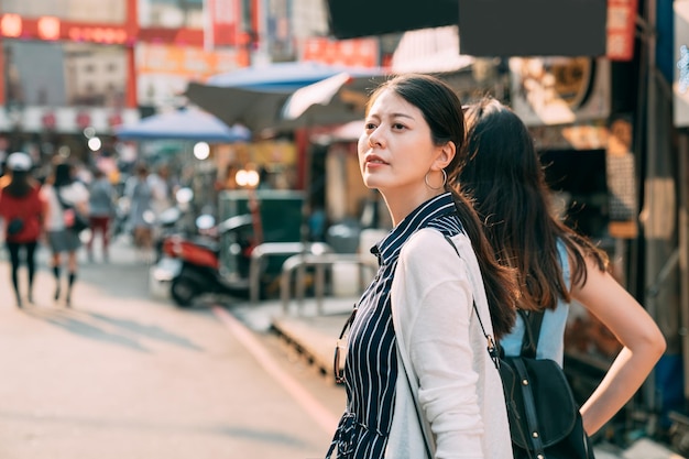 사진 아름다운 호기심 많은 아시아 여성이 햇살 아래 관광 비즈니스 지역의 거리에서 친구와 쇼핑을 하는 동안 거리를 보기 위해 돌아서고 있습니다.