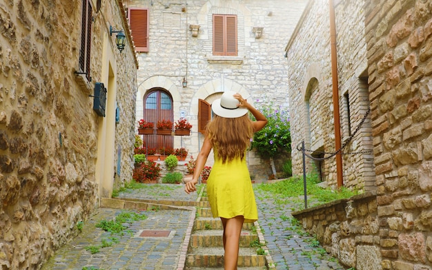 Beautiful curious young woman with yellow dress and hat goes upstairs in street in Assisi, Italy. Rear view of happy cheerful girl visiting central Italy.