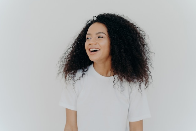 Beautiful curly adult woman turns gaze aside has glad expression smiles and laughs at something funny