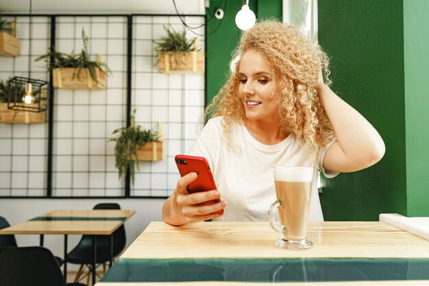 Beautiful curly blonde woman taking selfie while sitting at the table in coffee shop