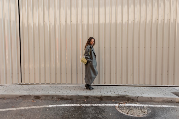 Bella ragazza riccia alla moda in abiti vintage di moda con una borsa cammina vicino a un muro di metallo. lo street style e la bellezza delle donne