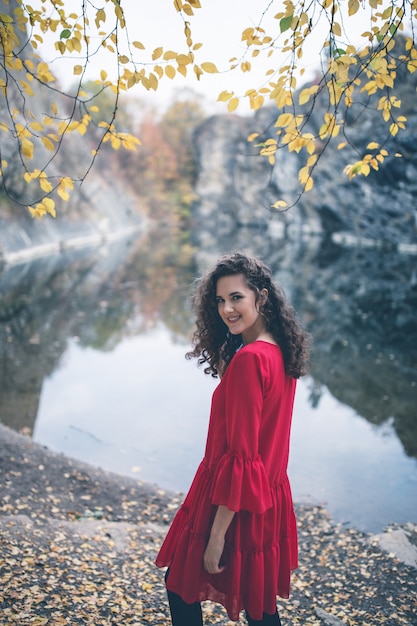 Красивая девушка вьющиеся волосы в красном платье, стоя на берегу озера и улыбается