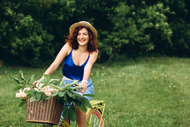 Bella ragazza dai capelli ricci in un cappello sorriso e tiene in bicicletta con cesto di fiori