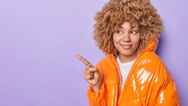 Beautiful curly haired woman points left shows something\
dressed in orange jacket demonstrates promotion or place for your\
advertisement isolated over purple background look at this promo\
banner