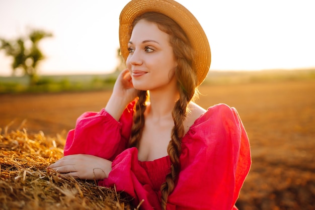 日没時に田舎の干し草の俵の近くでポーズをとる帽子と服を着た美しい巻き毛の女性