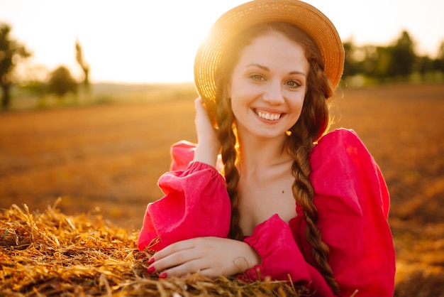 Красивая кудрявая женщина в шляпе и одежде позирует возле тюков сена в сельской местности на закате