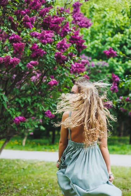 Una bella donna riccia in un abito estivo corre e si rallegra di indossare occhiali da sole sullo sfondo di un cespuglio di lillà viola