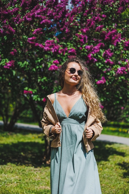 여름 드레스를 입은 아름다운 곱슬머리 여성이 보라색 라일락 덤불의 배경에 선글라스를 끼고 달리고 기뻐합니다