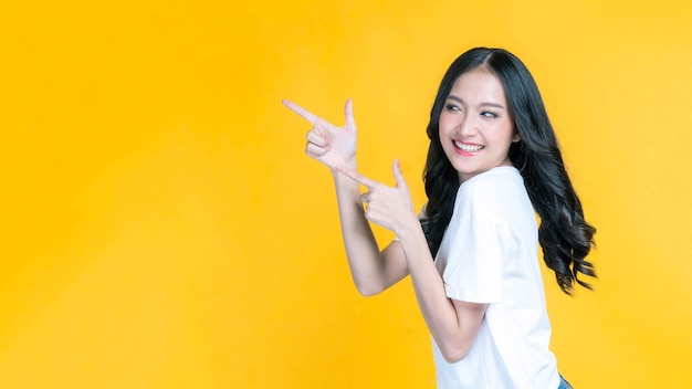 写真 美しい可愛いアジア人女性が笑顔で指を指して空のコピースペースを示しています 黄色い背景に隔離された広告のための空白スペース