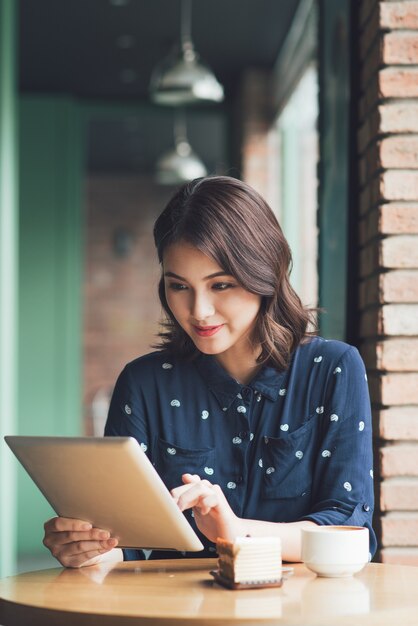 Красивая милая азиатская молодая коммерсантка в кафе, используя цифровой планшет и пьет кофе улыбается
