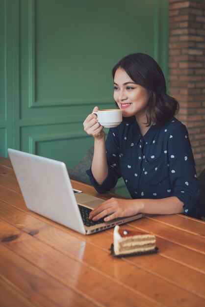 ノートパソコンを使用して、笑顔でコーヒーを飲みながら、カフェで美しいかわいいアジアの若い実業家