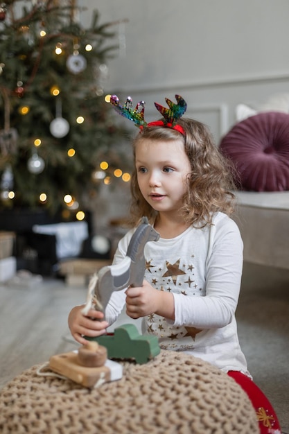 Beautiful cute child girl in fashionable christmas clothes sits near the christmas tree and plays with a wooden horse on holidays