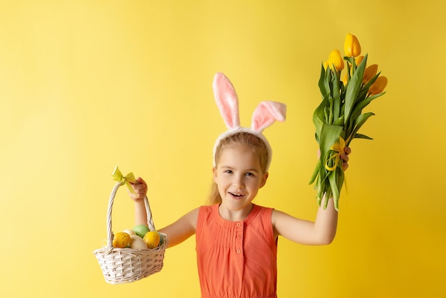 부활절 토끼 귀에 아름다운 귀여운 소녀는 노란색 배경에 계란 바구니를 들고