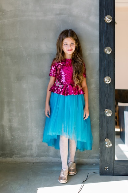 Beautiful cute little girl in a pink shiny blouse and a blue skirt on a grey background standing near mirror.