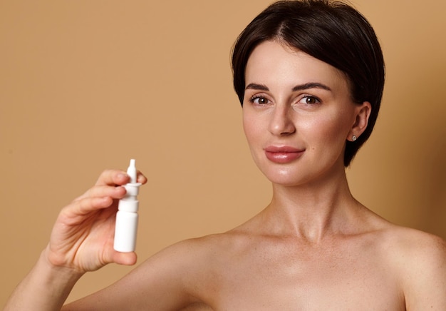 Beautiful cute smiling Caucasian woman cute smiles looking at camera, holding nasal spray isolated on beige background with copy ad space. Antiallergic and symptomatic treatment of rhinitis