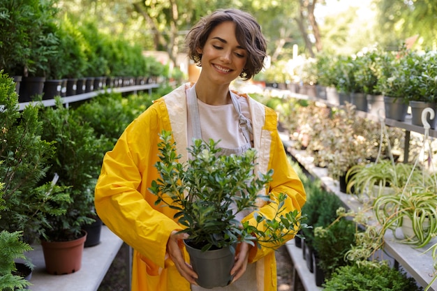 植物を保持している温室で花の植物の上に立っている美しいかわいい女性の庭師