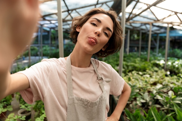 beautiful cute woman gardener standing over plants in greenhouse take a selfie showing tongue.