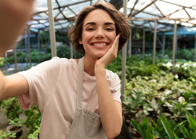 温室の植物の上に立っている美しいかわいい女性の庭師は、自分撮りを取ります。