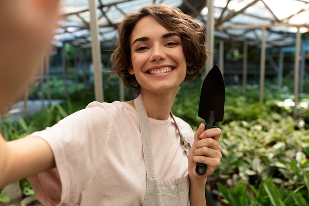 Bella donna carina giardiniere in piedi sopra le piante in serra prendere un selfie.