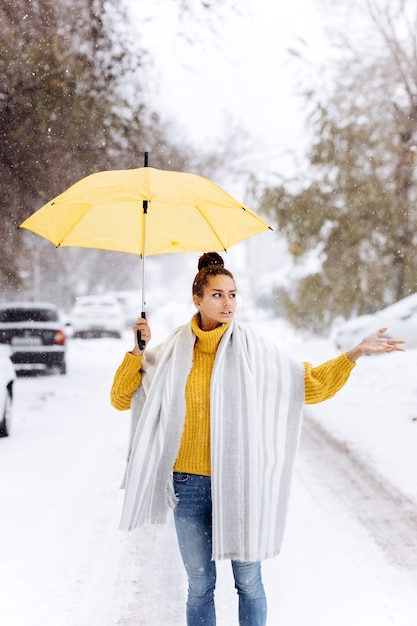 Красивая темноволосая девушка, одетая в желтый свитер, джинсы и белый шарф, стоит с желтым зонтиком на заснеженной улице в зимний день.
