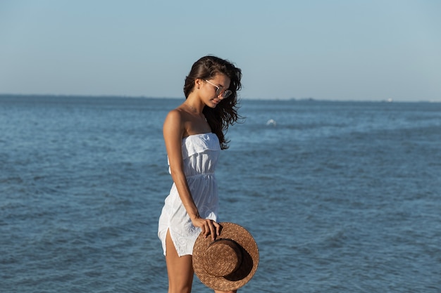 Photo beautiful dark-haired girl in white dress and sunglasses hold hat in hand and walk near the sea on a sunny day. .