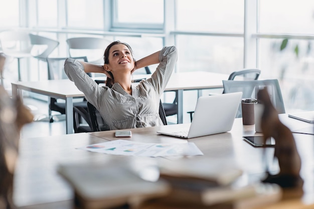 Beautiful dark-haired woman dreaming with opened eyes in office chair. Businesswoman feels relaxed after busy working day. Female leaning back in chair with hands behind head. Satisfied girl resting a