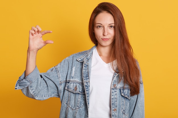 beautiful dark haired woman wears demin jacket shows something small with hand, isolated against yellow wall