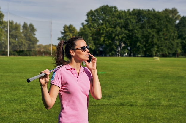 Красивая темноволосая молодая кавказская гольфистка в круглых очках звонит по мобильному телефону на открытом воздухе