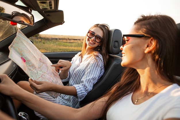 Beautiful dark-haired young women in sunglasses are sitting in a black cabriolet on a sunny day. One of them keeps map in her hands. .