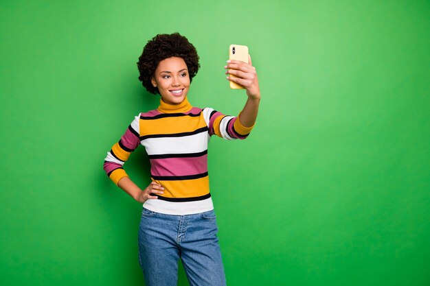 Di una bella signora riccia di pelle scura che tiene le mani al telefono facendo selfie per il popolare sito web online, indossa jeans pullover a righe casual