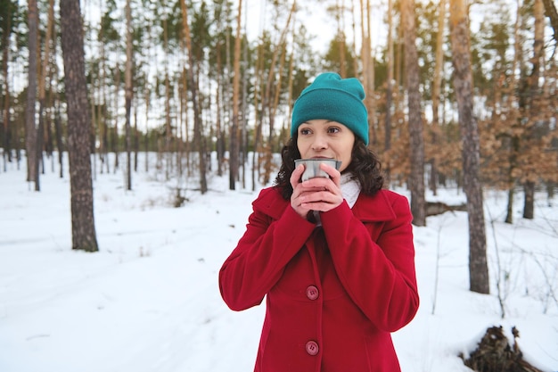 Beautiful darkhaired European pretty woman in bright red winter woolen coat and green knit hat drinking tea or coffee from a thermo mug resting in a snow covered forest enjoying a weekend outdoor