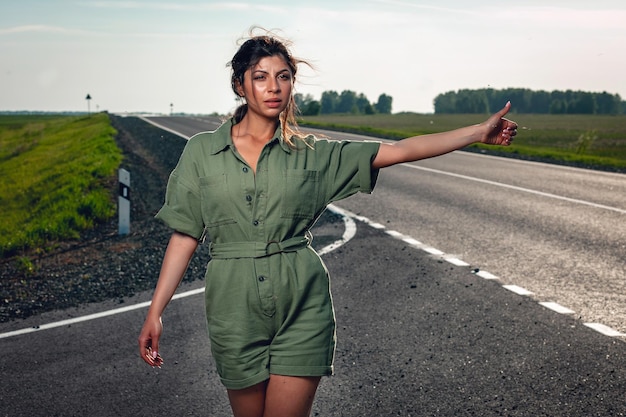 Beautiful disheveled sexy girl in a tight overalls is hitchhiking on the road, portrait