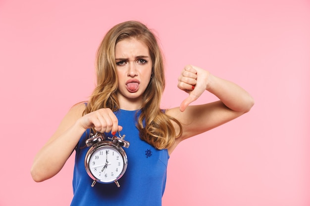 a beautiful displeased young pretty woman posing isolated over pink wall holding alarm clock