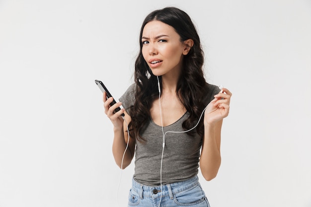 Una bella giovane donna dispiaciuta in posa isolata sul muro bianco ascoltando musica con gli auricolari utilizzando il telefono cellulare.