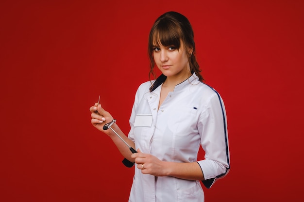 A beautiful doctor girl holds a reflex hammer and smiles at the camera isolated on a red background