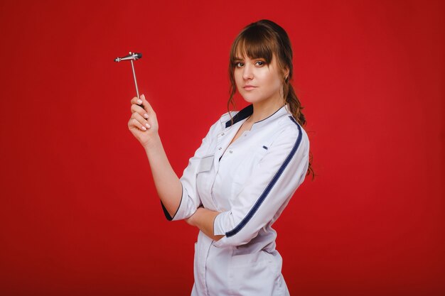 A beautiful doctor girl holds a reflex hammer and smiles at the camera isolated on a Red background.