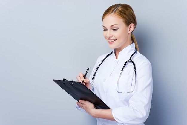 Beautiful doctor at work. Confident female doctor in white uniform writing in clipboard while standing against grey background