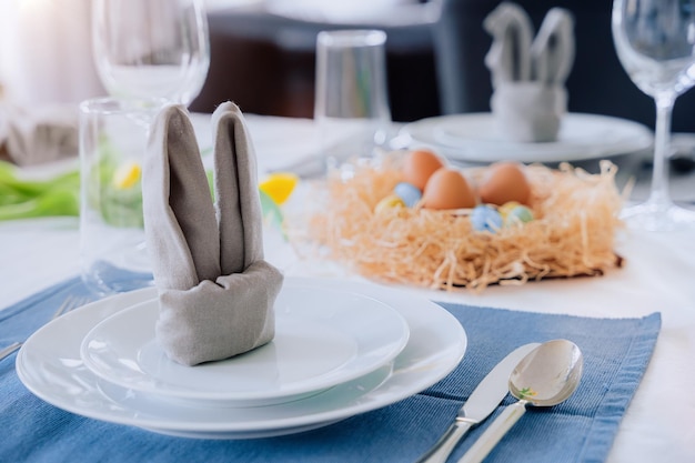 Beautiful Easter table setting with festive decor indoors Napkin in shape of rabbit ears colorful eggs in nest fresh yellow tulips in vase Sunlight postcard greeting Interior