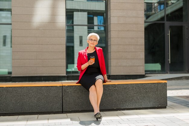 beautiful elderly woman in age with a short haircut and glasses in a red jacket with a cup of coffee