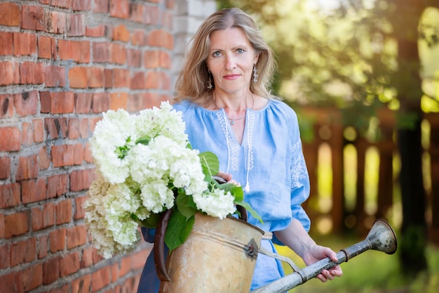 A beautiful elderly woman in an embroidered shirt holds a retro watering can with flowers